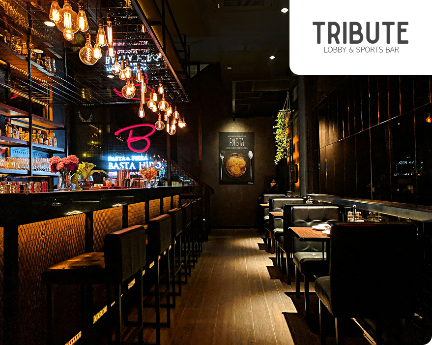 Tribute Lobby & Sports Bar, a nighttime-themed bar with bar stools, tables with armchairs, and numerous hanging light bulbs creating a cozy ambiance.